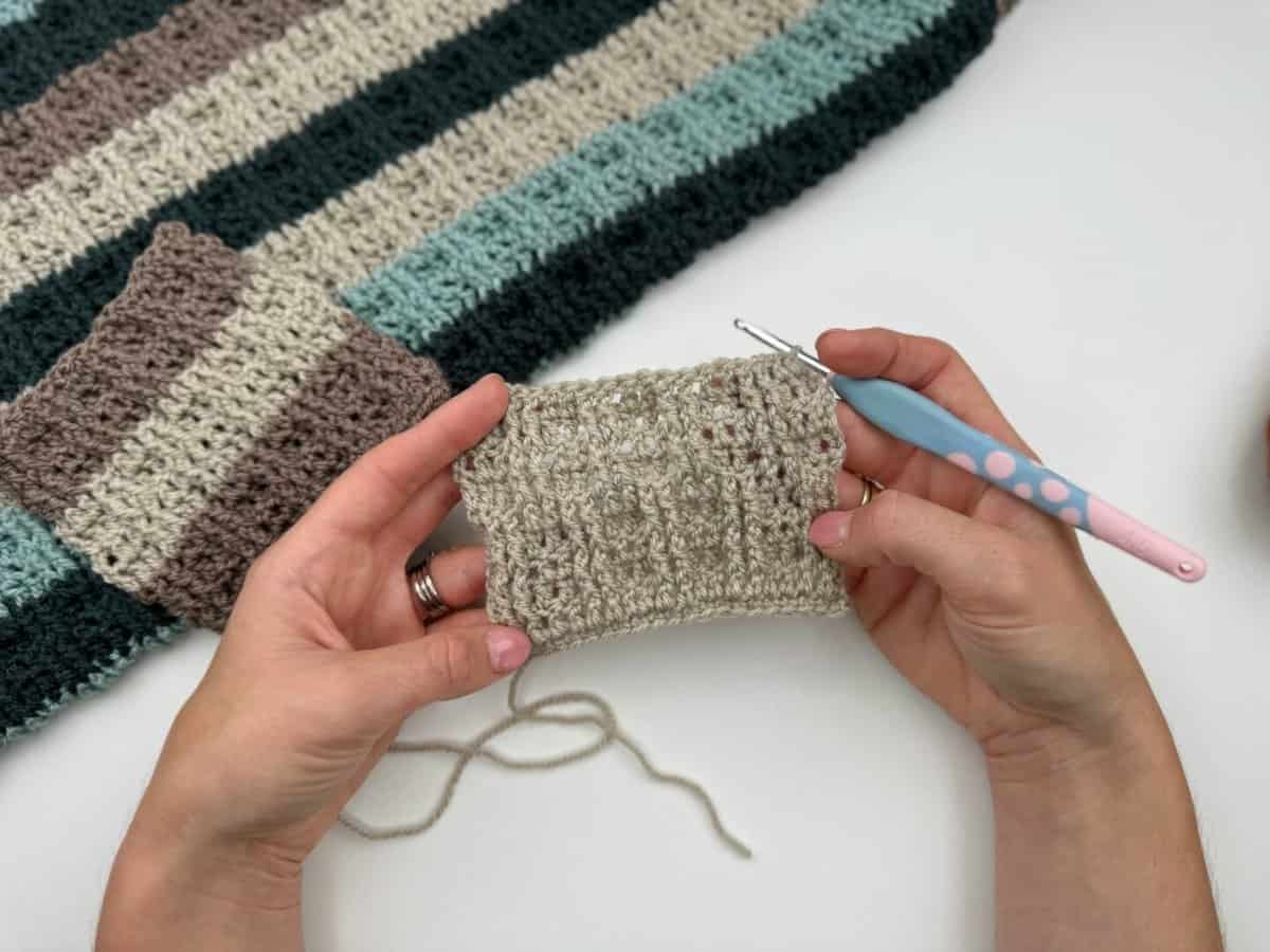A person holds a crochet swatch and hook, with a striped crochet project in beige, brown, and green on the table in the background, featuring alternating front and back post double crochet stitches.
