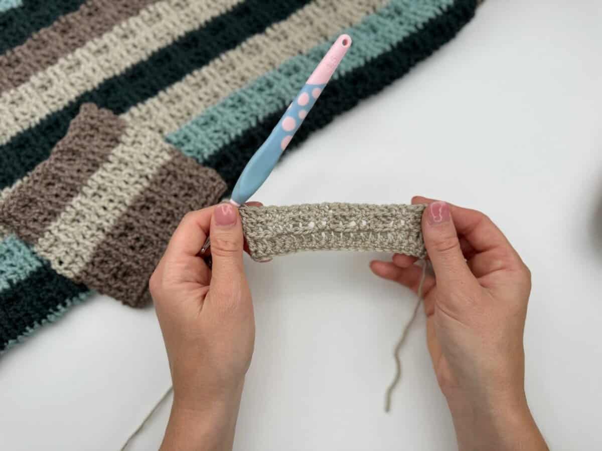 Hands holding a beige crochet swatch with a crochet hook, showcasing alternating front and back post double crochet; a striped crochet blanket is partially visible in the background.