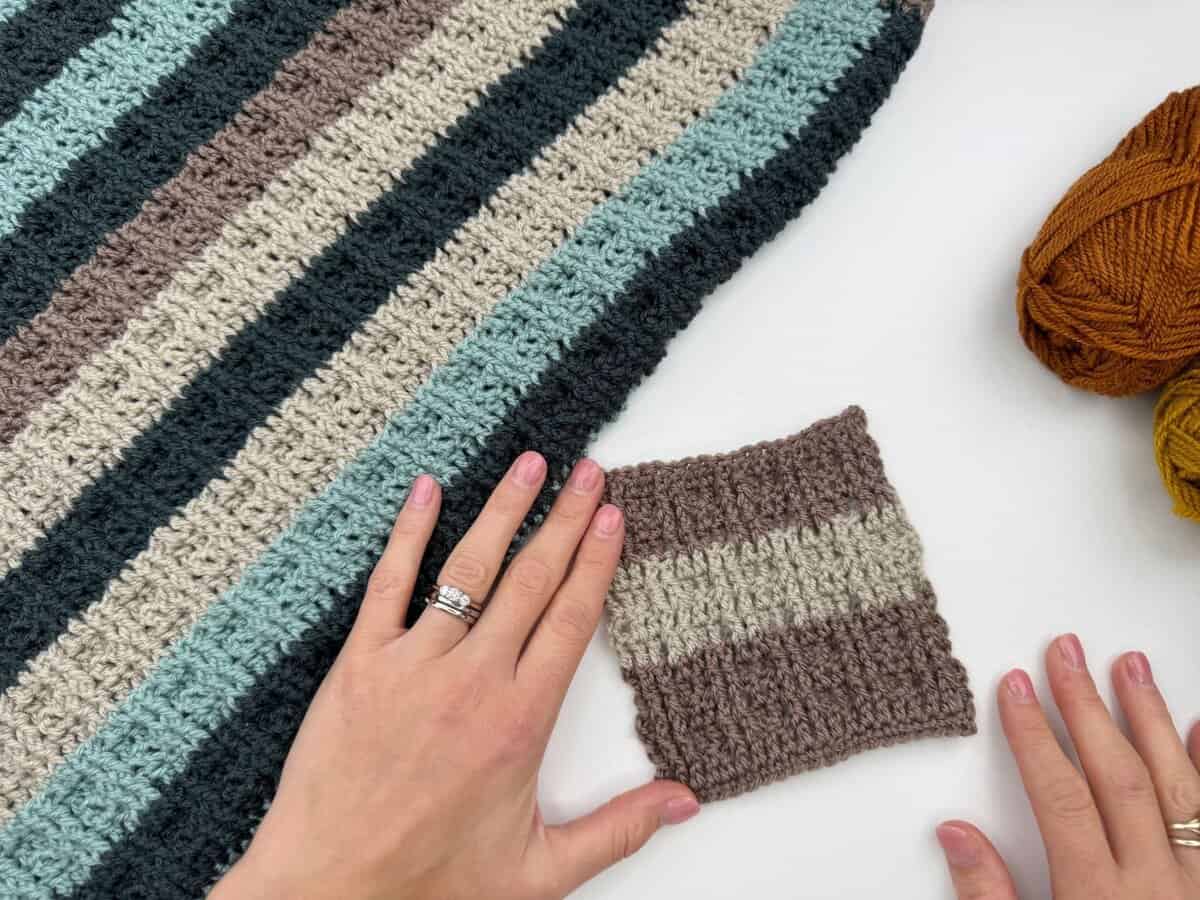 A hand holds a small crocheted swatch next to a larger striped crocheted piece, with skeins of yarn nearby on a white surface.