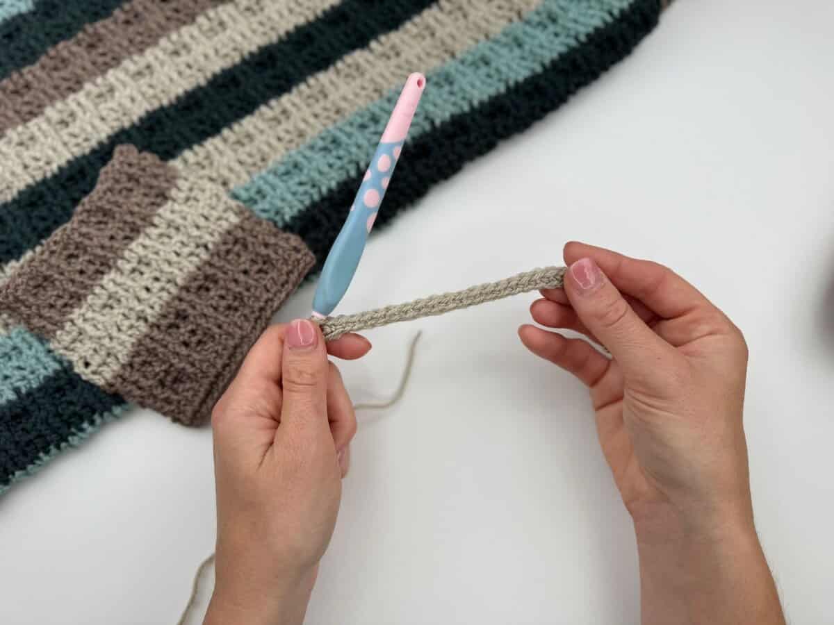 Hands are crocheting with a light blue and pink crochet hook, starting a row of beige yarn using alternating front and back post double crochet; a striped crocheted blanket and swatch are in the background.