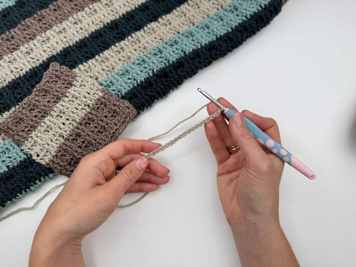 A person holds a crochet hook and yarn, starting a new chain next to a striped, partially finished project on a table, featuring alternating front and back post double crochet stitches.