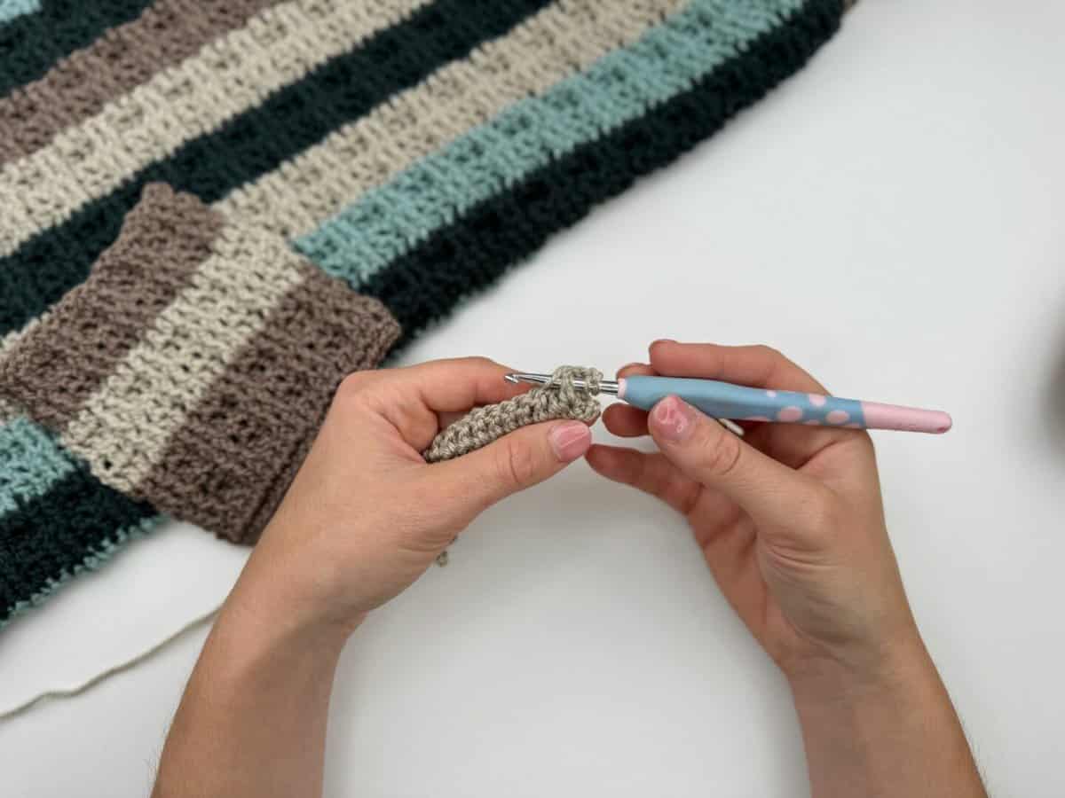 Hands holding a crochet hook and working with beige yarn in front of a striped crocheted fabric piece on a white surface, showcasing alternating front and back post double crochet stitches.