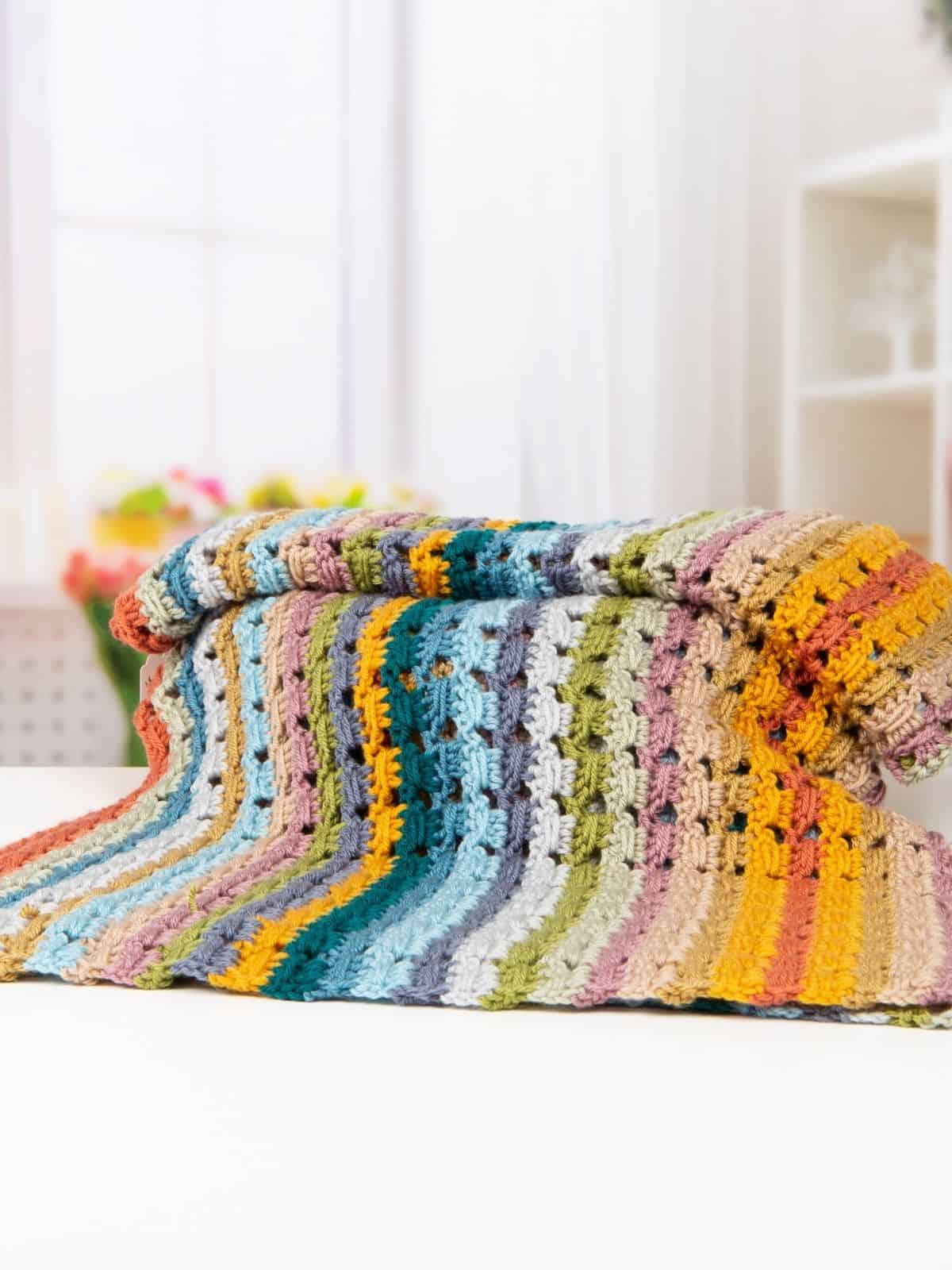 A folded, multicolored striped crochet blanket made from a simple crochet blanket pattern is displayed on a white surface with a softly focused background.