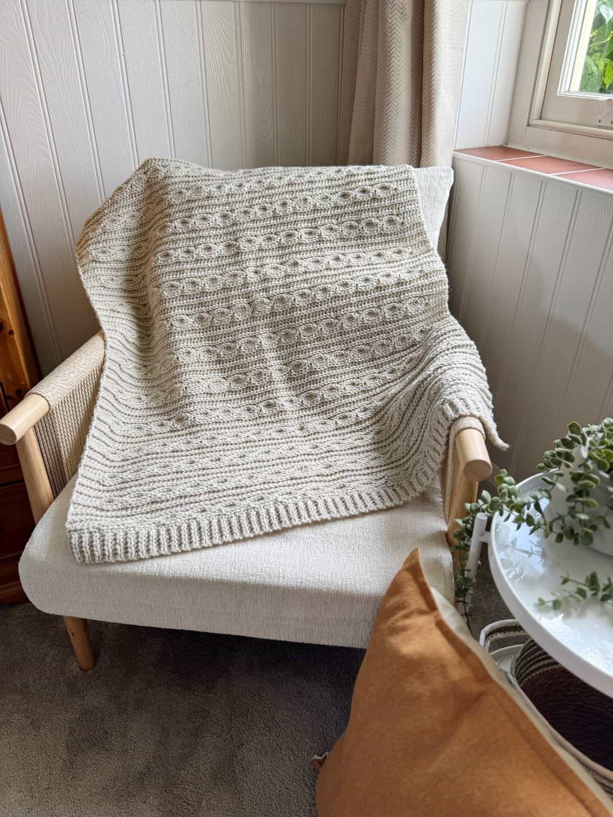 A textured beige crocheted blanket, crafted in a modern crochet blanket pattern, draped over a light-colored cushioned chair beside a white round side table with a plant and an orange cushion.