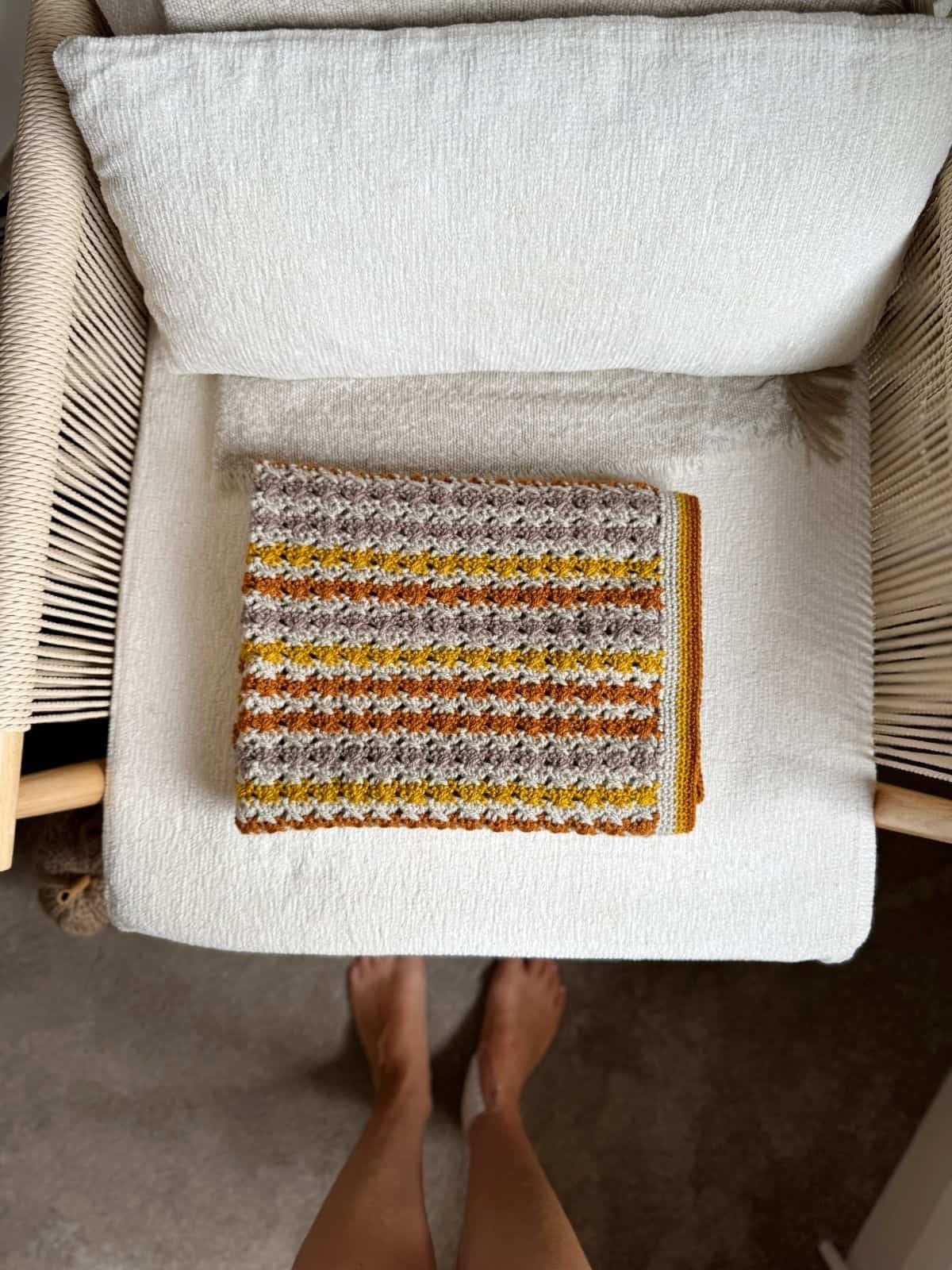 A folded, multicolored crochet blanket with yellow, brown, and gray stripes—perfect inspiration for a simple crochet blanket pattern—sits on a white cushioned chair; bare feet are visible on the floor in front of the chair.
