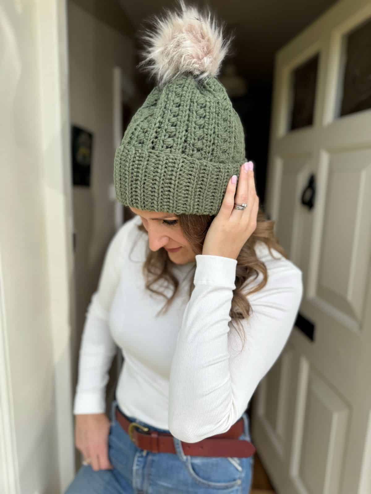 Woman wearing an olive green crochet beanie inspired by a puff stitch crochet hat pattern, with a faux fur pom-pom, white long-sleeve shirt, and blue jeans stands indoors by a white door, looking down and touching her hat.