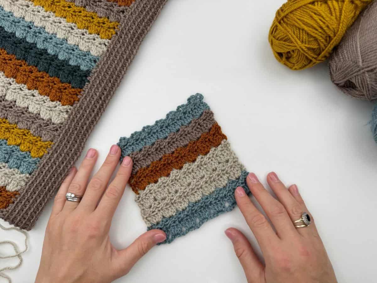 Two hands hold a small crochet swatch with horizontal stripes using the crochet cabbage patch stitch, next to a larger striped crochet piece and balls of yarn on a white surface.