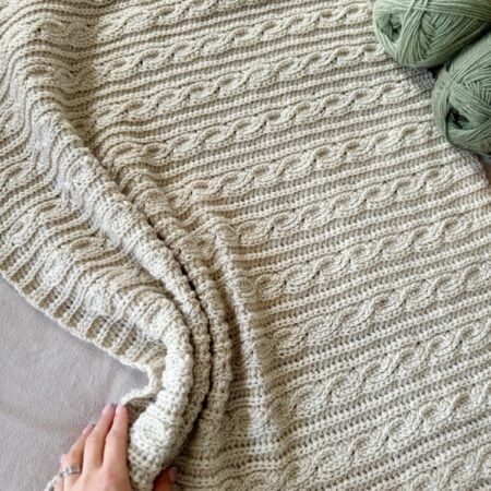A hand touches a textured, cream-colored crocheted blanket with a cable pattern. Two skeins of green yarn are placed nearby, offering inspiration for your next modern crochet blanket pattern.