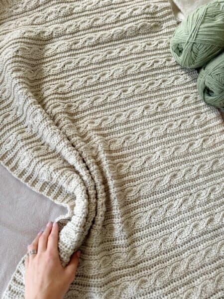 A hand touches a textured, cream-colored crocheted blanket with a cable pattern. Two skeins of green yarn are placed nearby, offering inspiration for your next modern crochet blanket pattern.