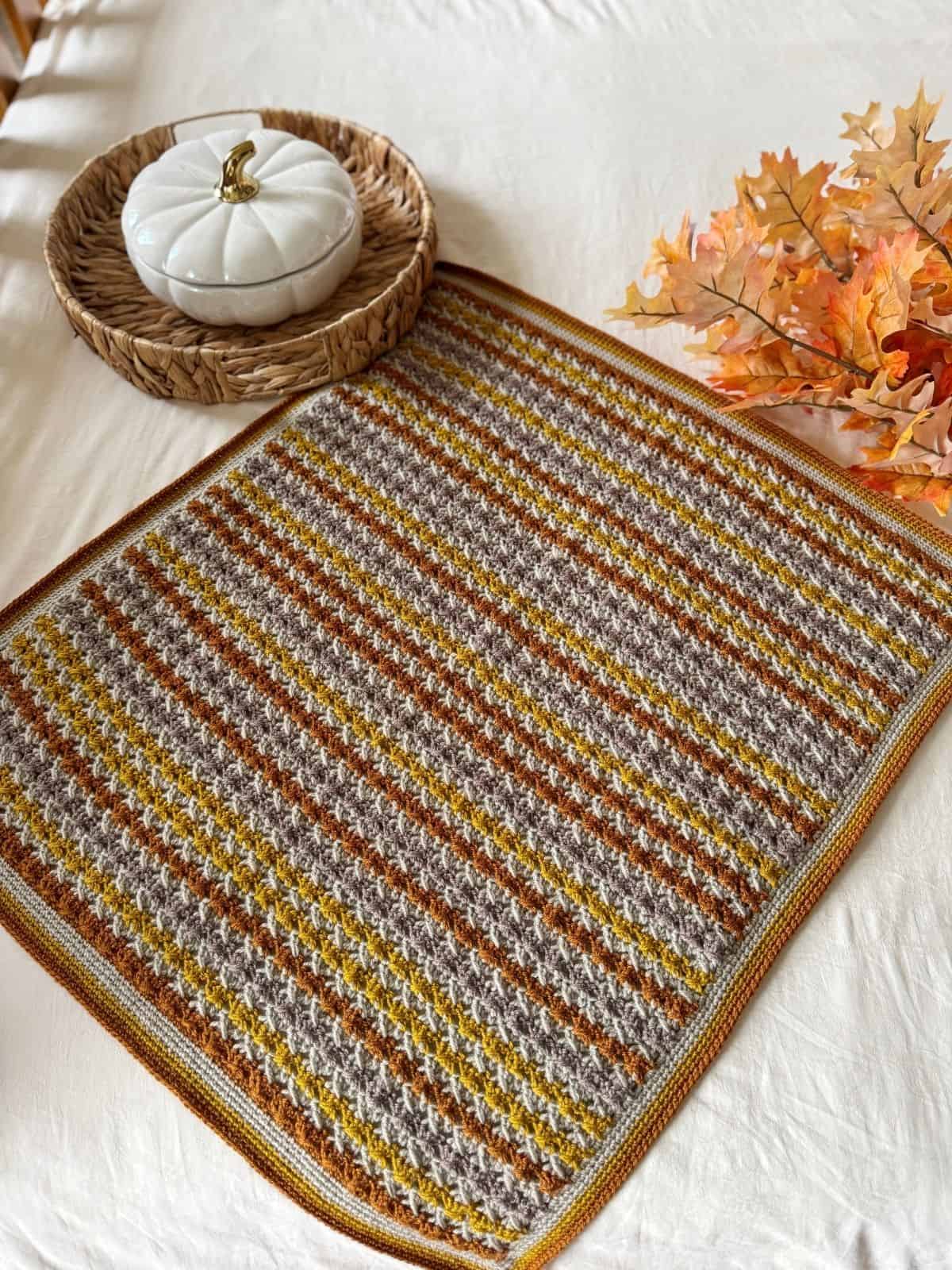 A simple crochet blanket pattern with brown, yellow, and gray stripes is laid on a table beside a wicker tray holding a white ceramic pumpkin and artificial autumn leaves.