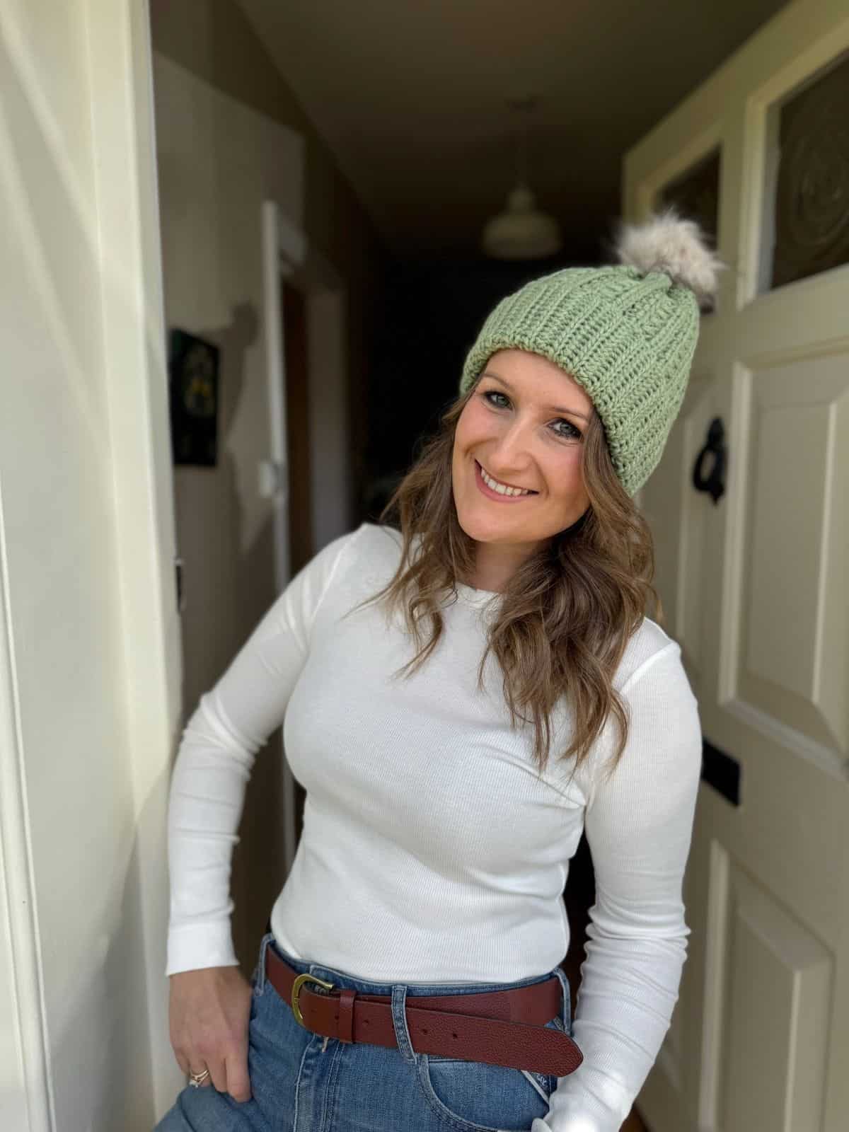 A woman wearing a light green puff stitch crochet hat, white long-sleeve shirt, and jeans stands smiling in a doorway with one hand in her pocket.
