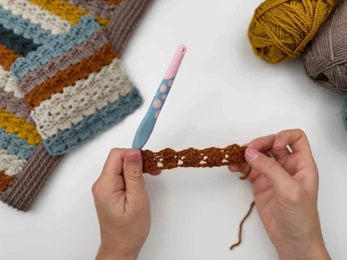 Two hands hold a brown crochet piece, worked in the cabbage patch stitch, with a pink and blue crochet hook; yarn balls and a crocheted fabric sample are nearby on a white surface.