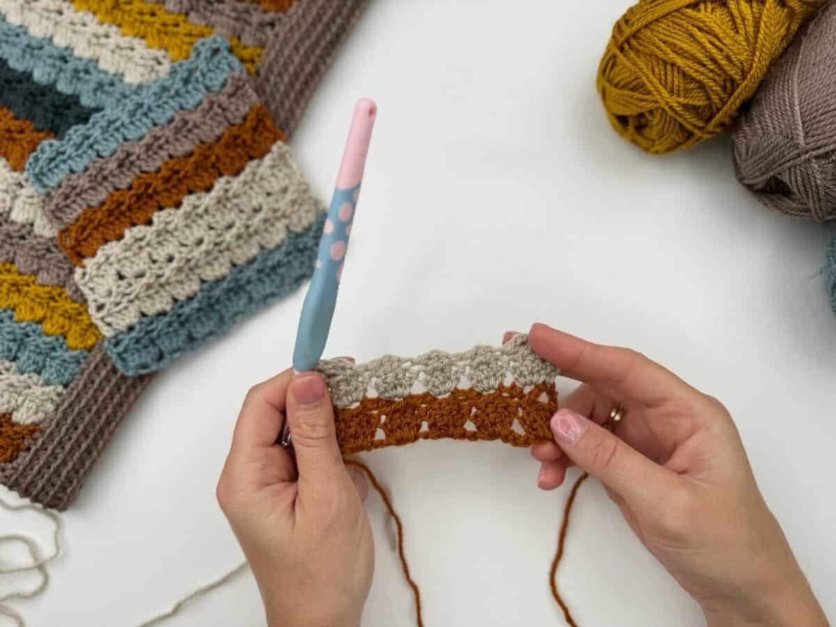 Hands holding a crochet project with a pink and blue crochet hook, multi-colored yarn, and a striped crochet cabbage patch stitch piece in the background.