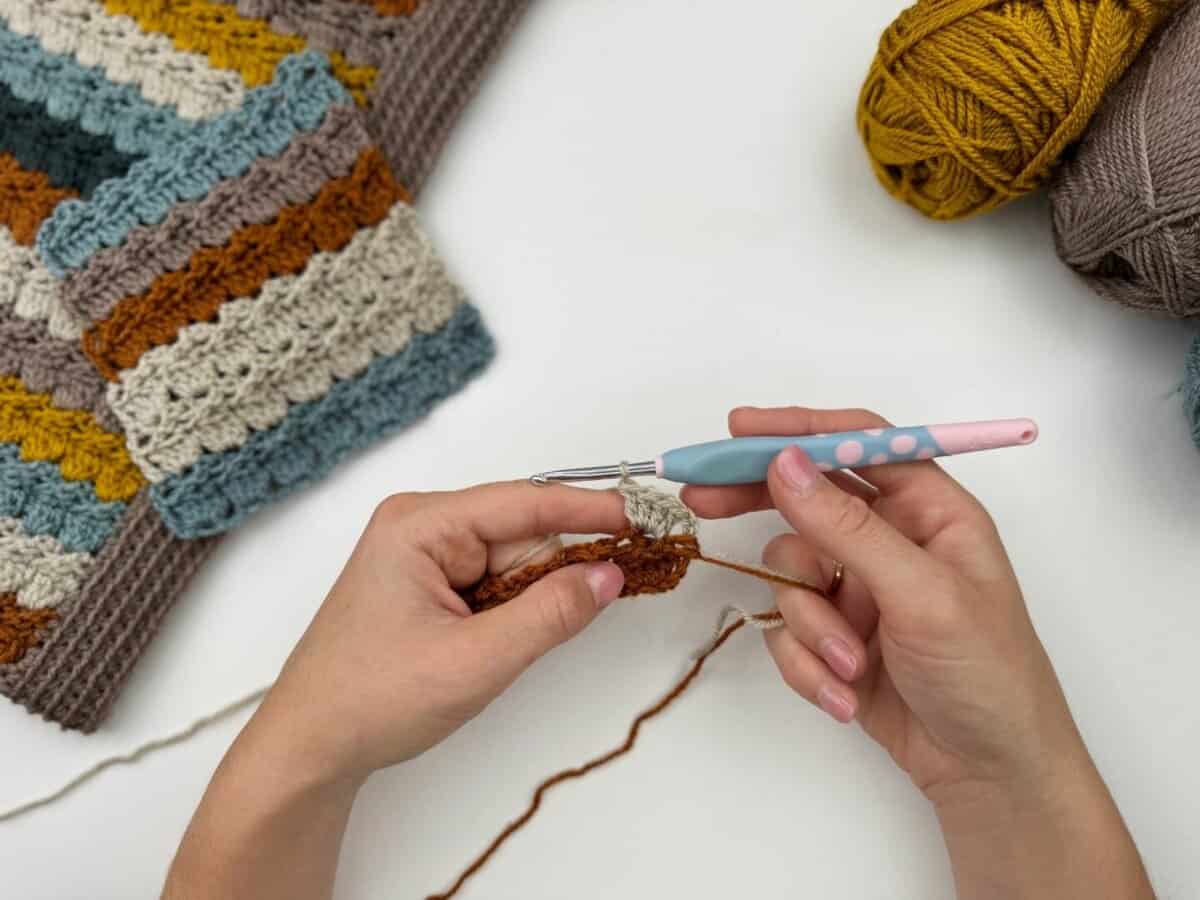 Hands crocheting with brown yarn using a crochet hook, showcasing the crochet cabbage patch stitch. Nearby are balls of yarn and a partially finished striped crochet piece on a white surface.