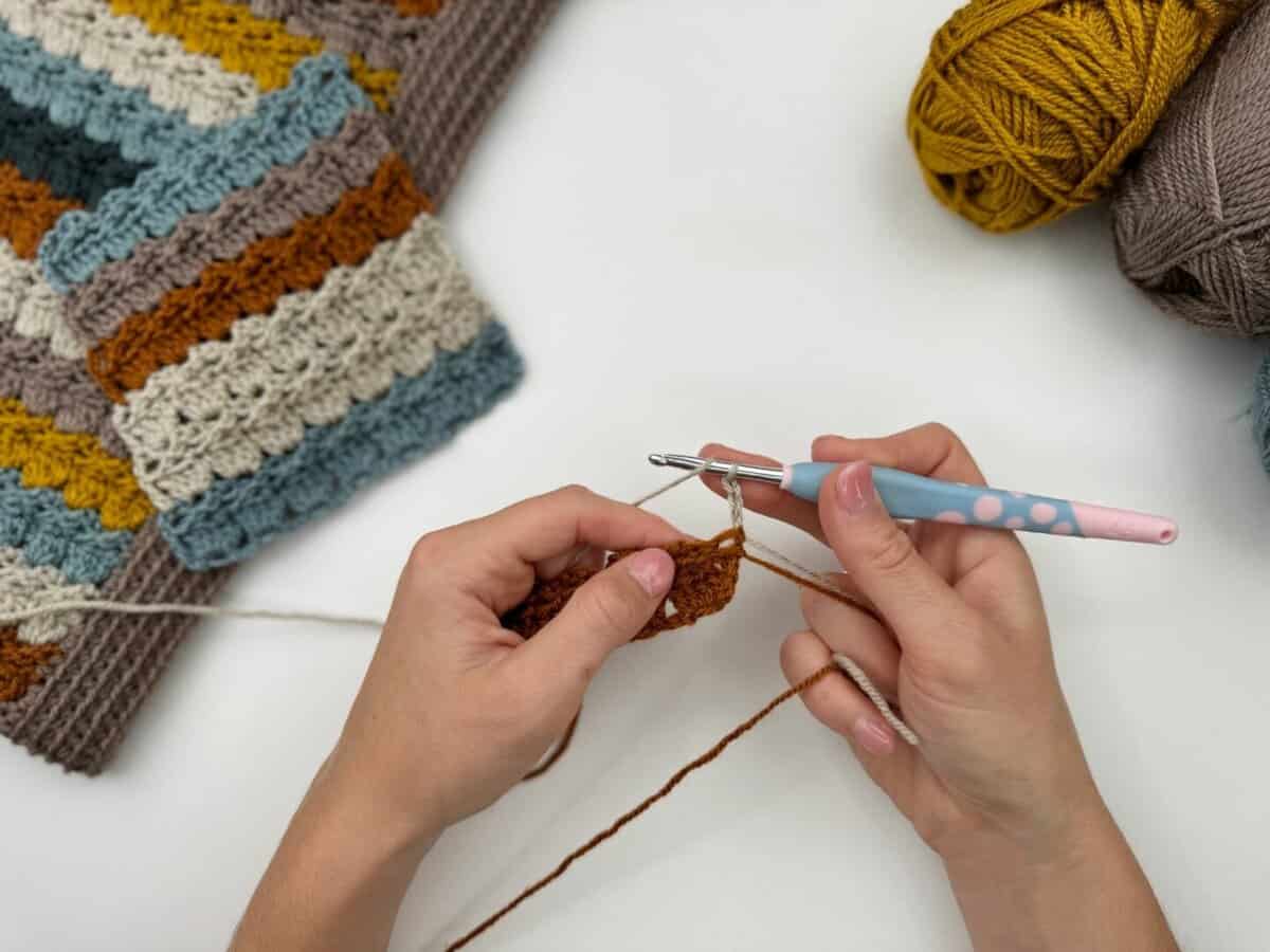 Hands crocheting with brown yarn and a pink crochet hook, using the crochet cabbage patch stitch, with balls of yarn and a multicolored crocheted piece on a white surface.
