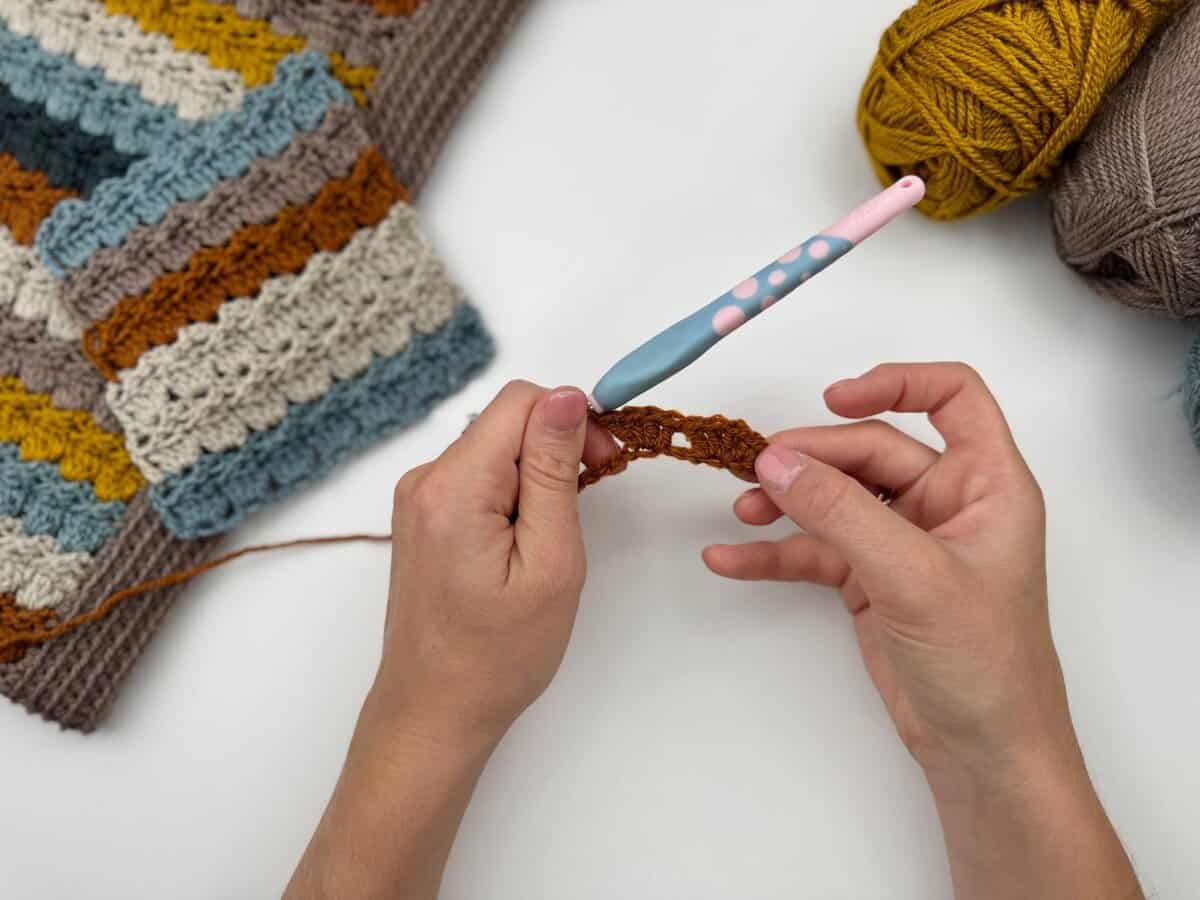 Two hands are crocheting with brown yarn and a pink and blue crochet hook, working the cabbage patch stitch. A striped crochet piece and balls of yellow and gray yarn rest nearby on a white surface.