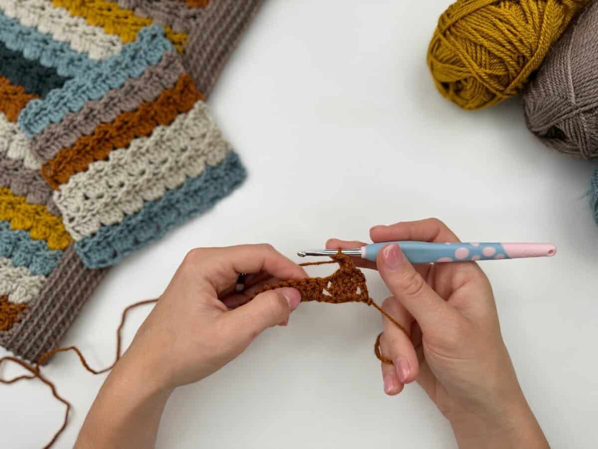 Two hands are crocheting with brown yarn and a pink crochet hook, working the crochet cabbage patch stitch. A striped crocheted piece and yarn balls rest on the white surface.
