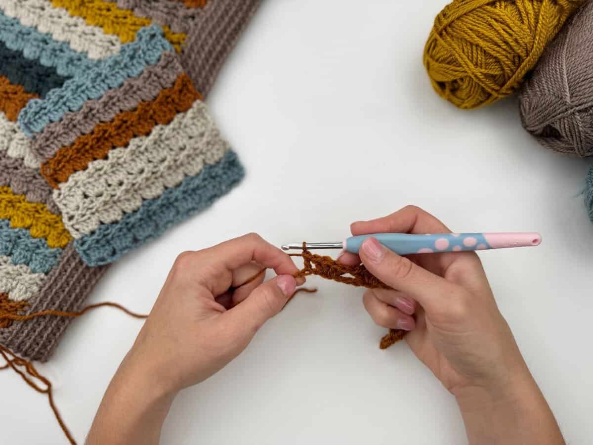 A person uses a crochet hook and brown yarn, working the crochet cabbage patch stitch, with crocheted pieces and two balls of yarn on a white surface.