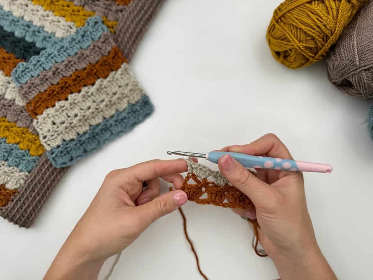Hands crocheting with a pink and blue crochet hook, surrounded by yarn balls and a striped crocheted piece on a white surface, showcasing the intricate crochet cabbage patch stitch.