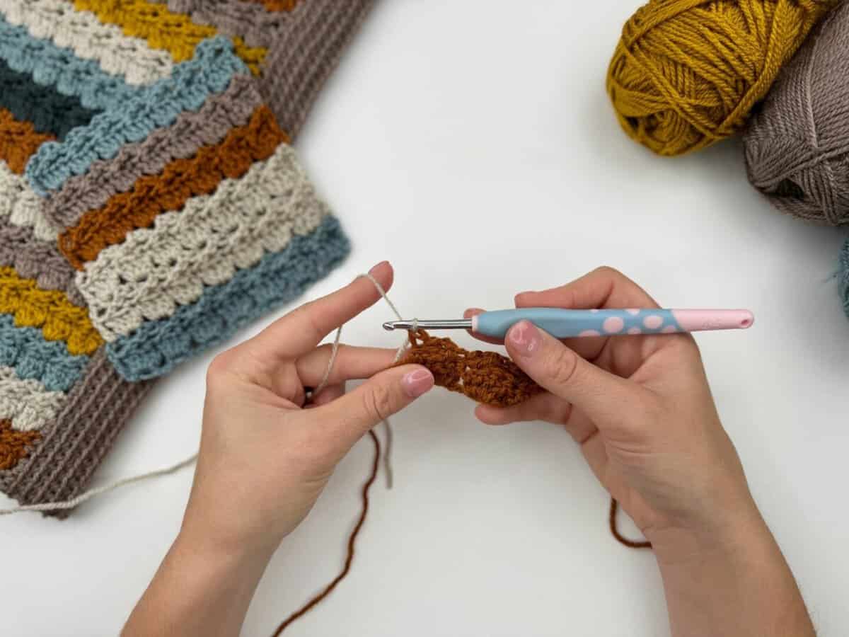 Two hands crochet cabbage patch stitch with a pink and blue hook using brown yarn, surrounded by striped crochet pieces and balls of yarn on a white surface.