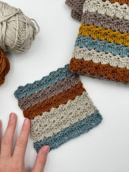 A hand touches a crocheted swatch featuring the crochet cabbage patch stitch with blue, beige, orange, brown, and yellow stripes, next to matching yarn balls and a larger crocheted piece on a white surface.