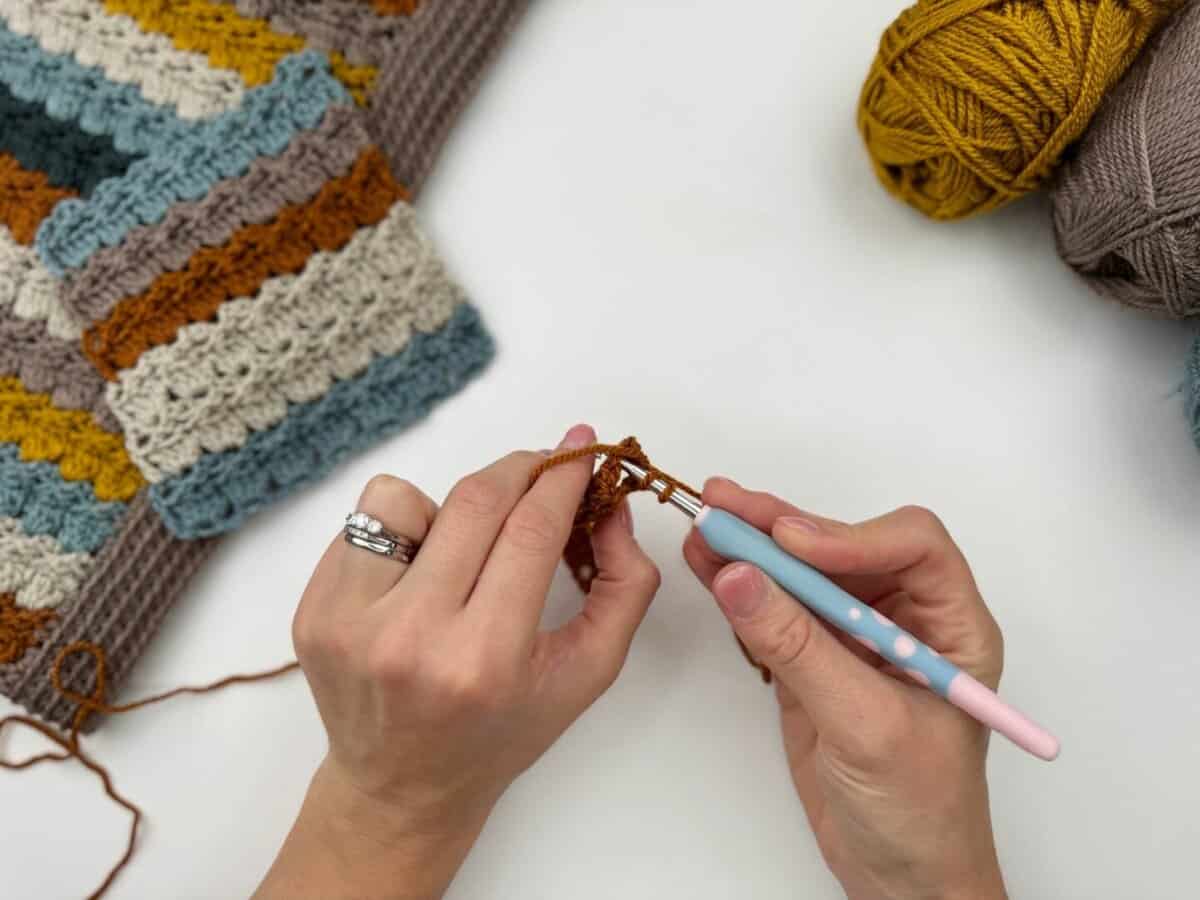 A person crochets with brown yarn using a blue crochet hook, working the cabbage patch stitch; finished multicolored crochet pieces and balls of yarn rest on a white surface.