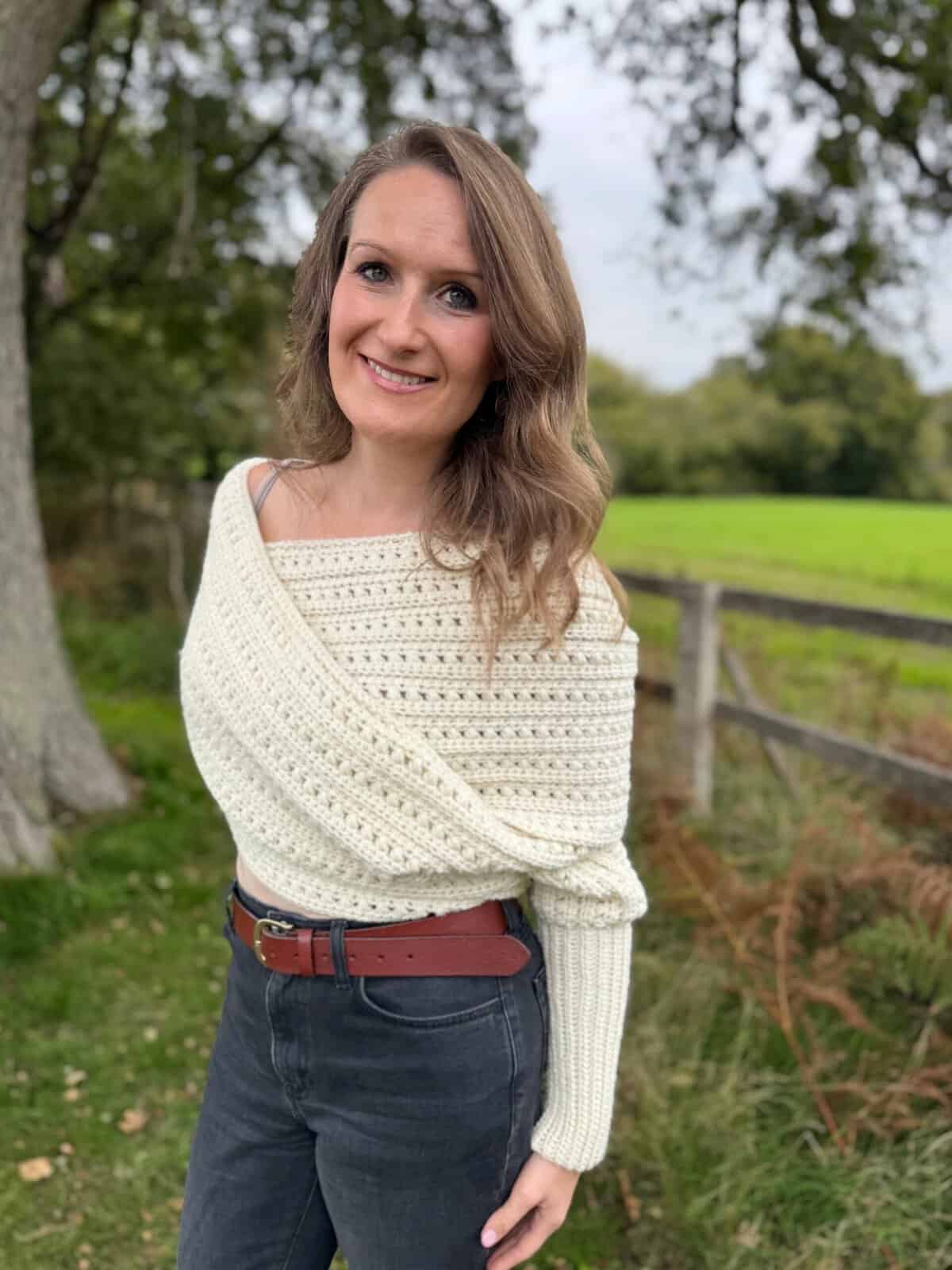 Woman standing outdoors near a wooden fence, wearing a cream knit sweater and black jeans with a brown belt, smiling at the camera. She completes her look with a cozy puff stitch crochet hat. Green grass and trees in the background.