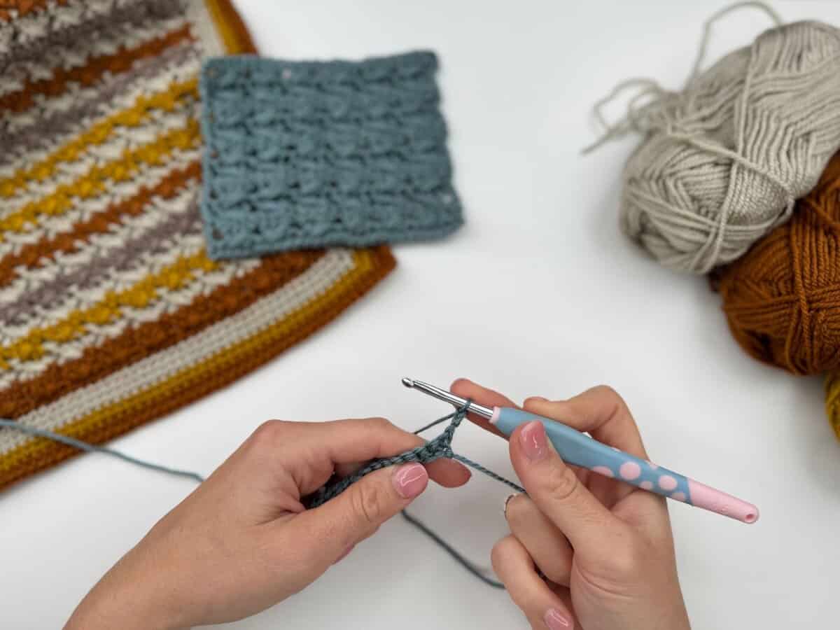 Hands crocheting with a blue and pink crochet hook, beside yarn balls and swatches of crocheted fabric on a white surface, perfect for following a Crossover Stitch Crochet Tutorial.