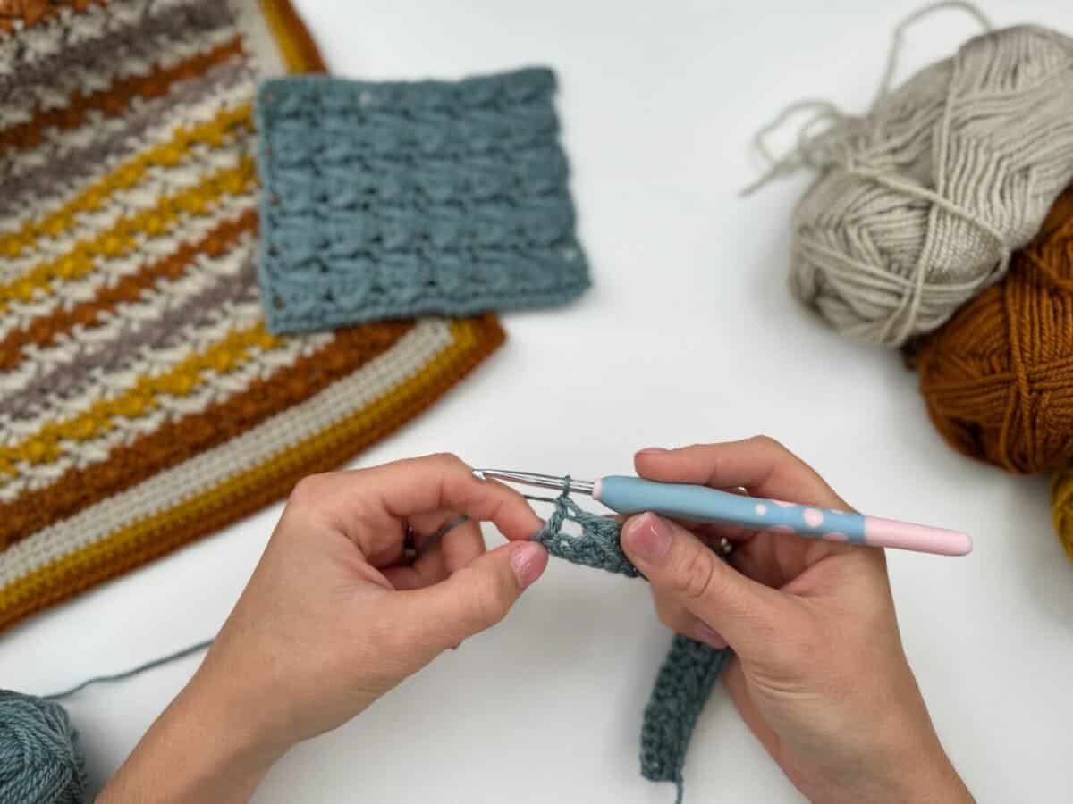 Hands crocheting with a blue-handled hook, working near finished crochet pieces and balls of yarn in cream, brown, and gold on a white surface—perfect for following along with a Crossover Stitch Crochet Tutorial.
