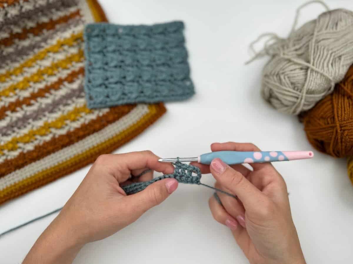 Close-up of hands working on a Crossover Stitch Crochet Tutorial with a pink and blue crochet hook; yarn balls and two finished crochet swatches rest on a white surface.