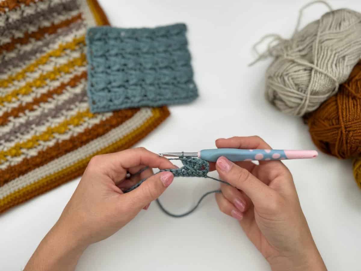 Two hands crochet with a blue hook and gray yarn; nearby are finished crochet squares and balls of brown, gray, and yellow yarn—a perfect setup for a Crossover Stitch Crochet Tutorial.