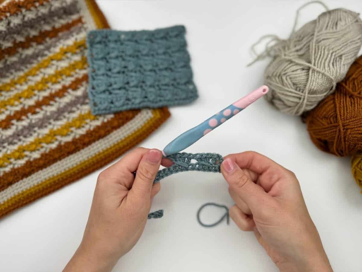 Hands crocheting with a blue hook and grey yarn, demonstrating the Crossover Stitch Crochet Tutorial, with finished crochet pieces and balls of yarn in brown, grey, and beige on a white surface.