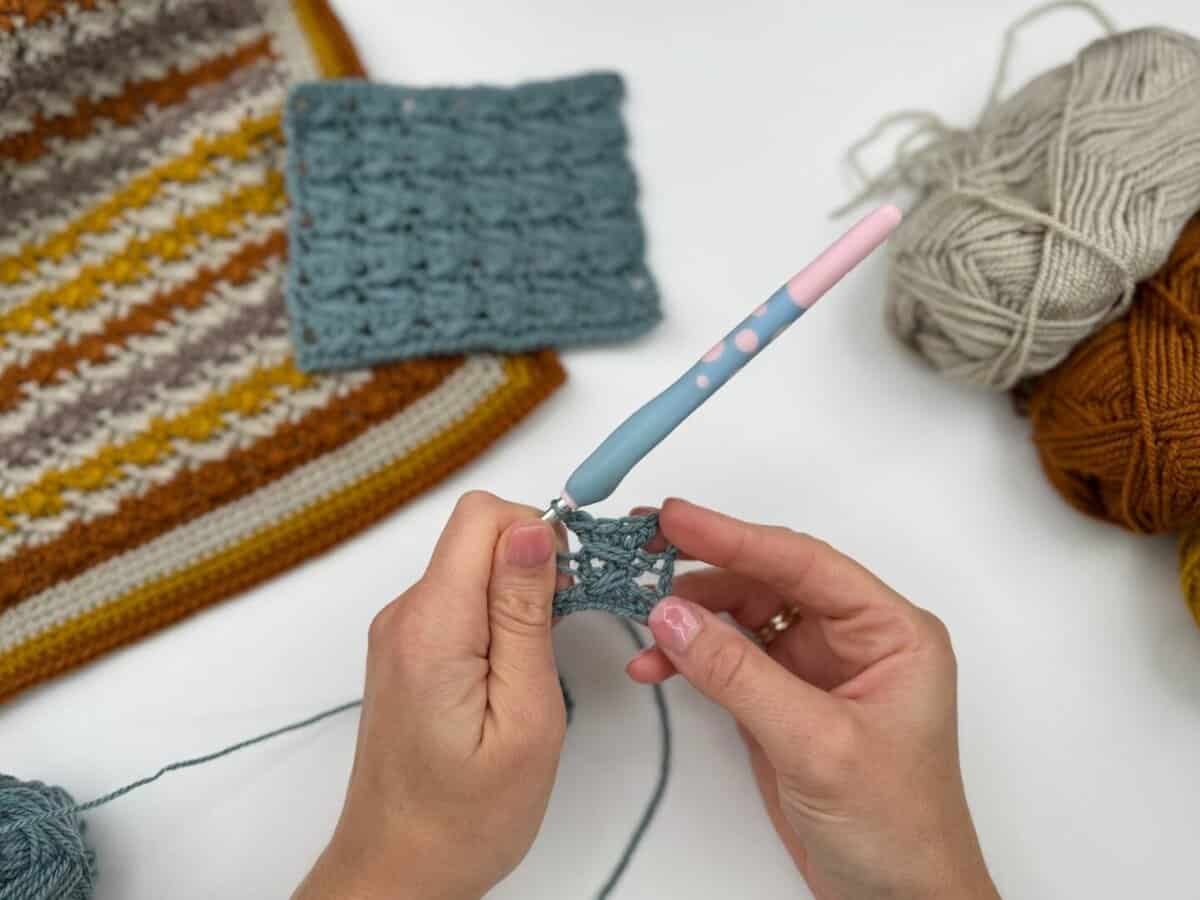 Close-up of hands working a Crossover Stitch Crochet Tutorial with blue yarn and a pink-and-blue crochet hook, surrounded by finished swatches and balls of yarn in the background.