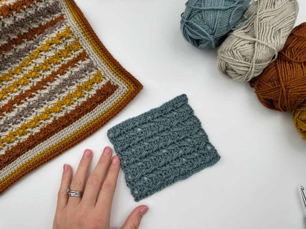 A hand rests near a blue crochet swatch, a striped crochet piece, and balls of yarn in blue, cream, and brown tones on a white surface—perfect for your next Crossover Stitch Crochet Tutorial.