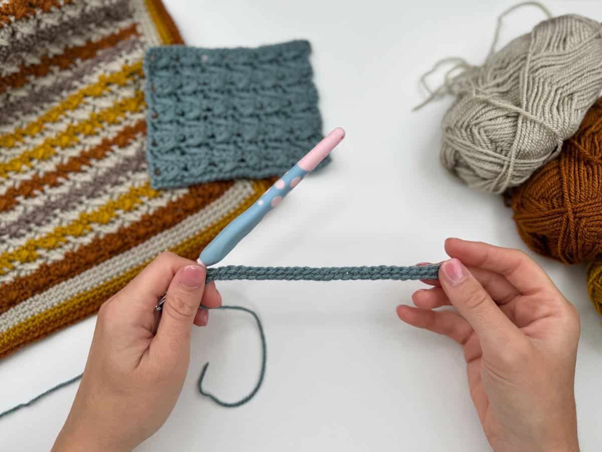 Two hands hold a crochet chain with a hook, ready for the Crossover Stitch Crochet Tutorial. Nearby are yarn balls, a crocheted blue swatch, and a striped crochet piece on a white surface.