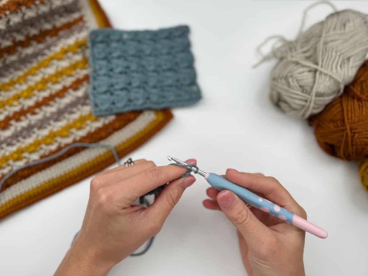 Hands crocheting with a blue and pink crochet hook, surrounded by yarn balls and two finished crochet swatches on a white surface—a perfect setup for following a Crossover Stitch Crochet Tutorial.