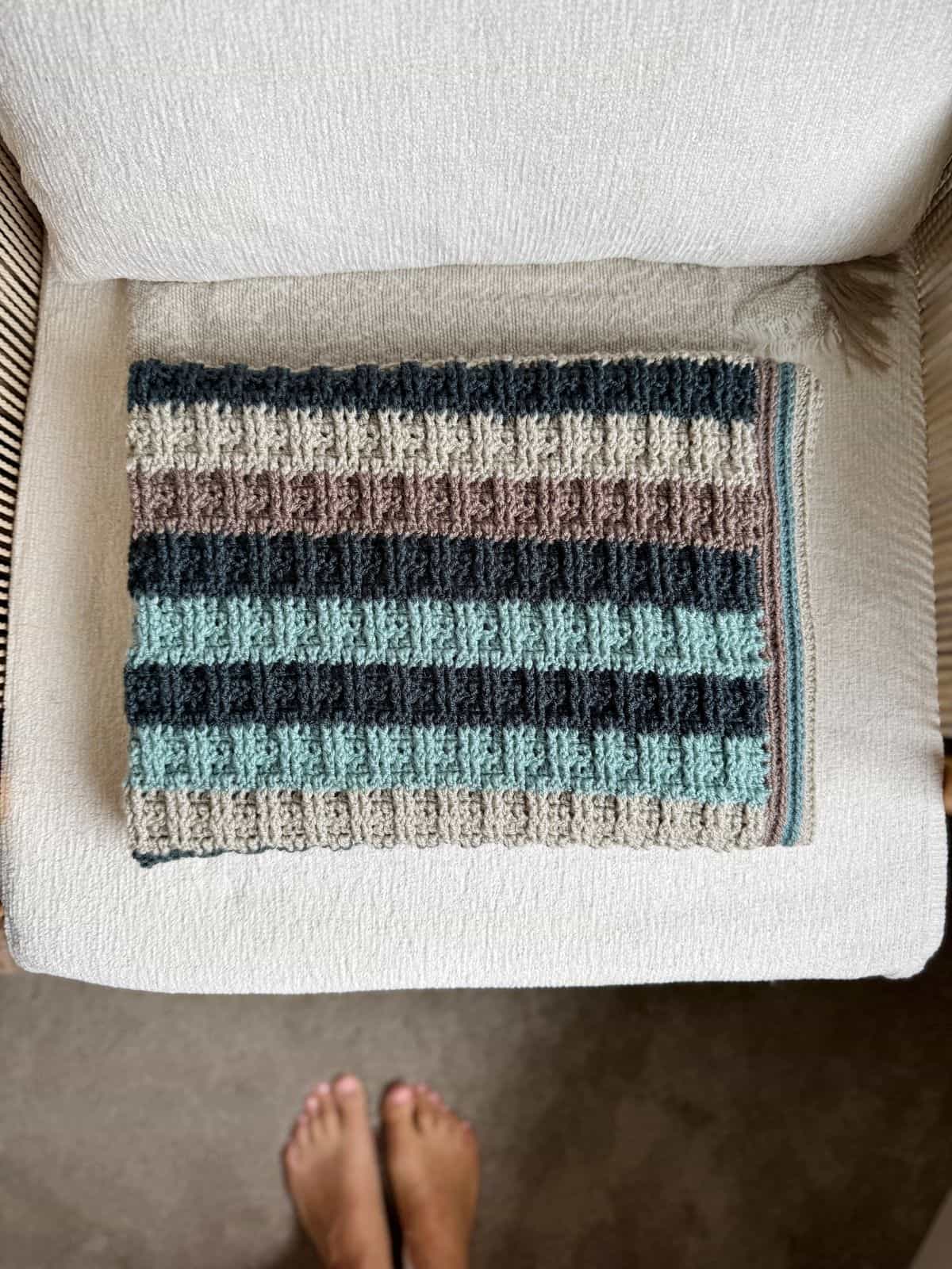 A folded, striped crochet blanket for boys in neutral colors rests on a light-colored upholstered chair, with bare feet visible at the bottom edge of the image.