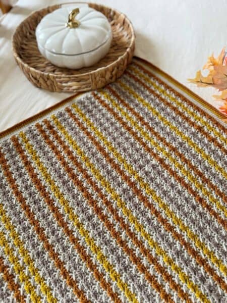A textured crochet blanket with brown, yellow, and cream stripes sits on a table next to a woven tray holding a white pumpkin-shaped container—an inviting scene inspired by a simple crochet blanket pattern.