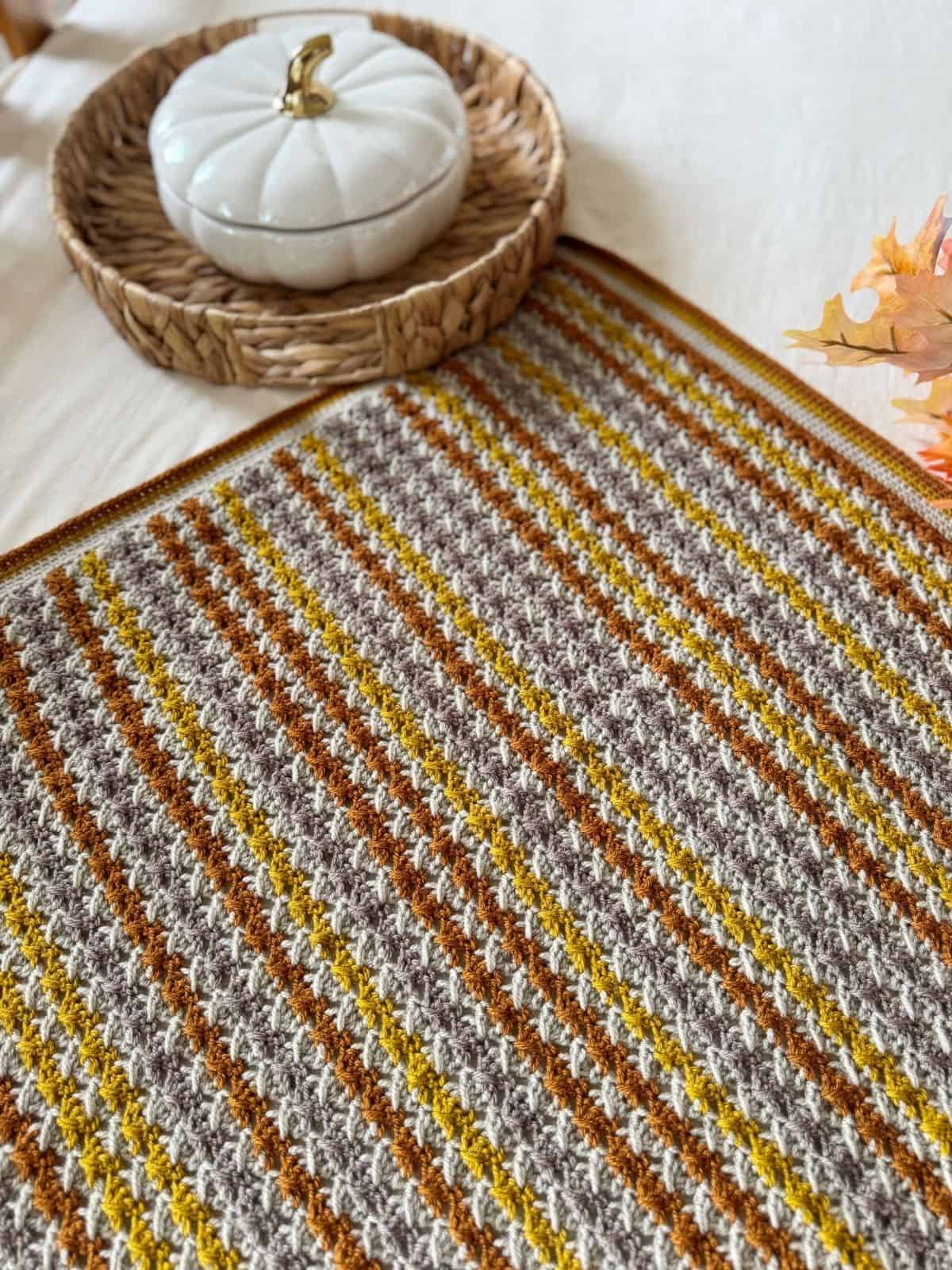 A textured crochet blanket with brown, yellow, and cream stripes sits on a table next to a woven tray holding a white pumpkin-shaped container—an inviting scene inspired by a simple crochet blanket pattern.