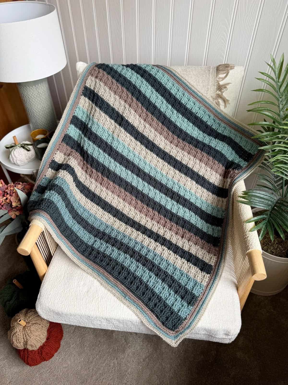 A striped crochet blanket in shades of teal, black, beige, and brown draped over a cream armchair beside a plant and a lamp.