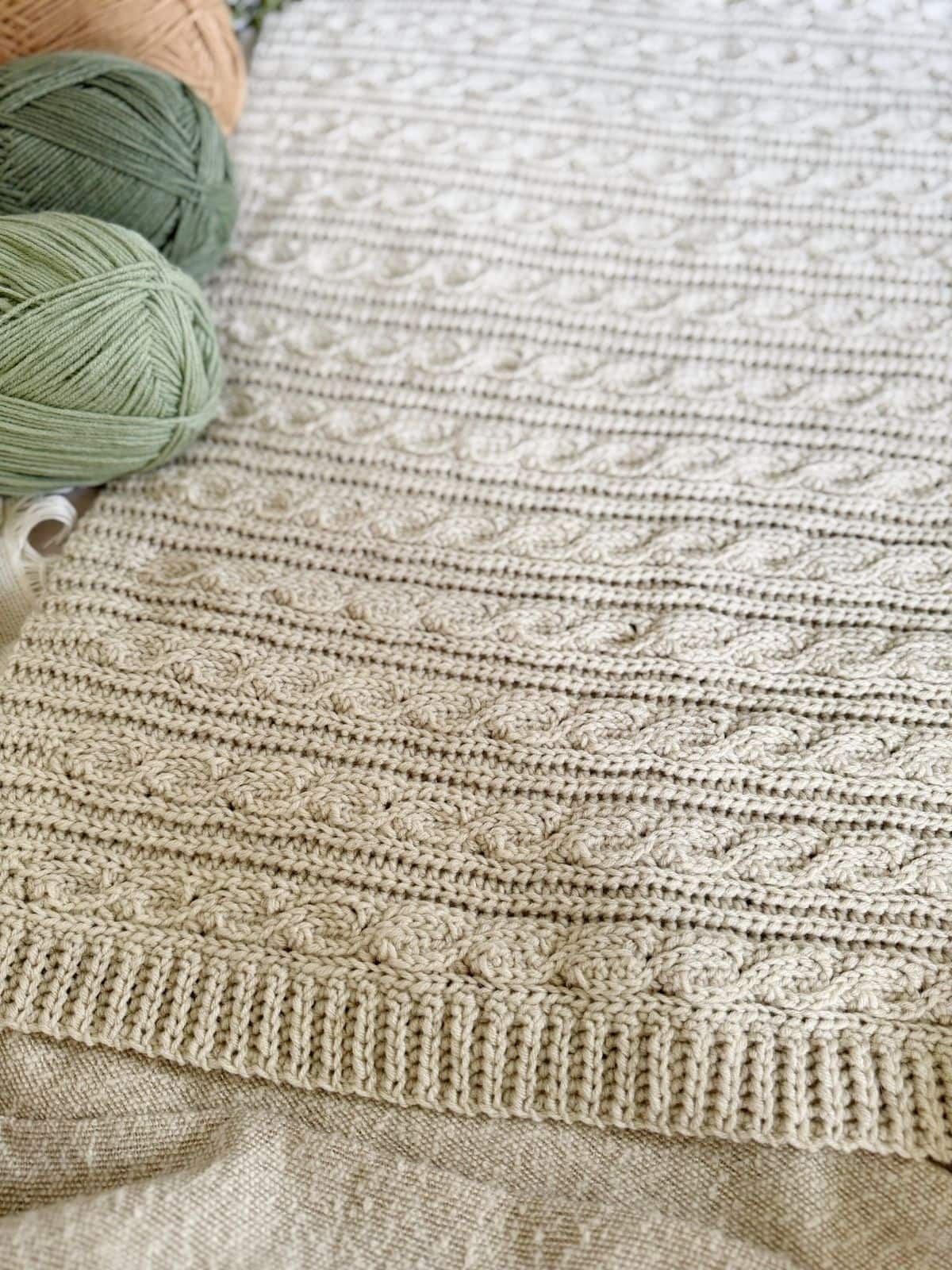Close-up of a beige crocheted fabric with a textured cable pattern, next to green and tan balls of yarn—perfect inspiration for a modern crochet blanket pattern.