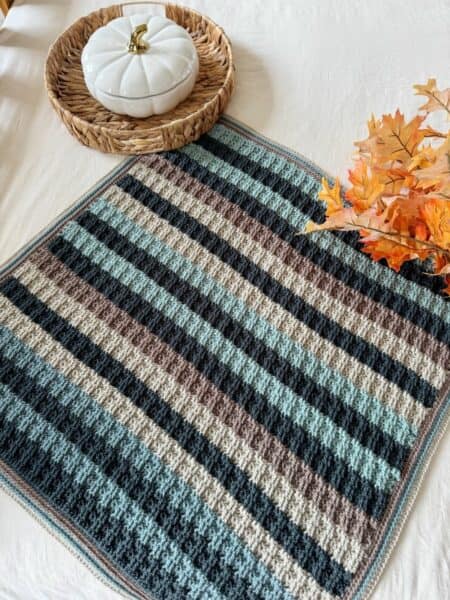 A crocheted striped square blanket in shades of blue, gray, and brown is displayed on a table with a ceramic pumpkin, wicker tray, and artificial autumn leaves.