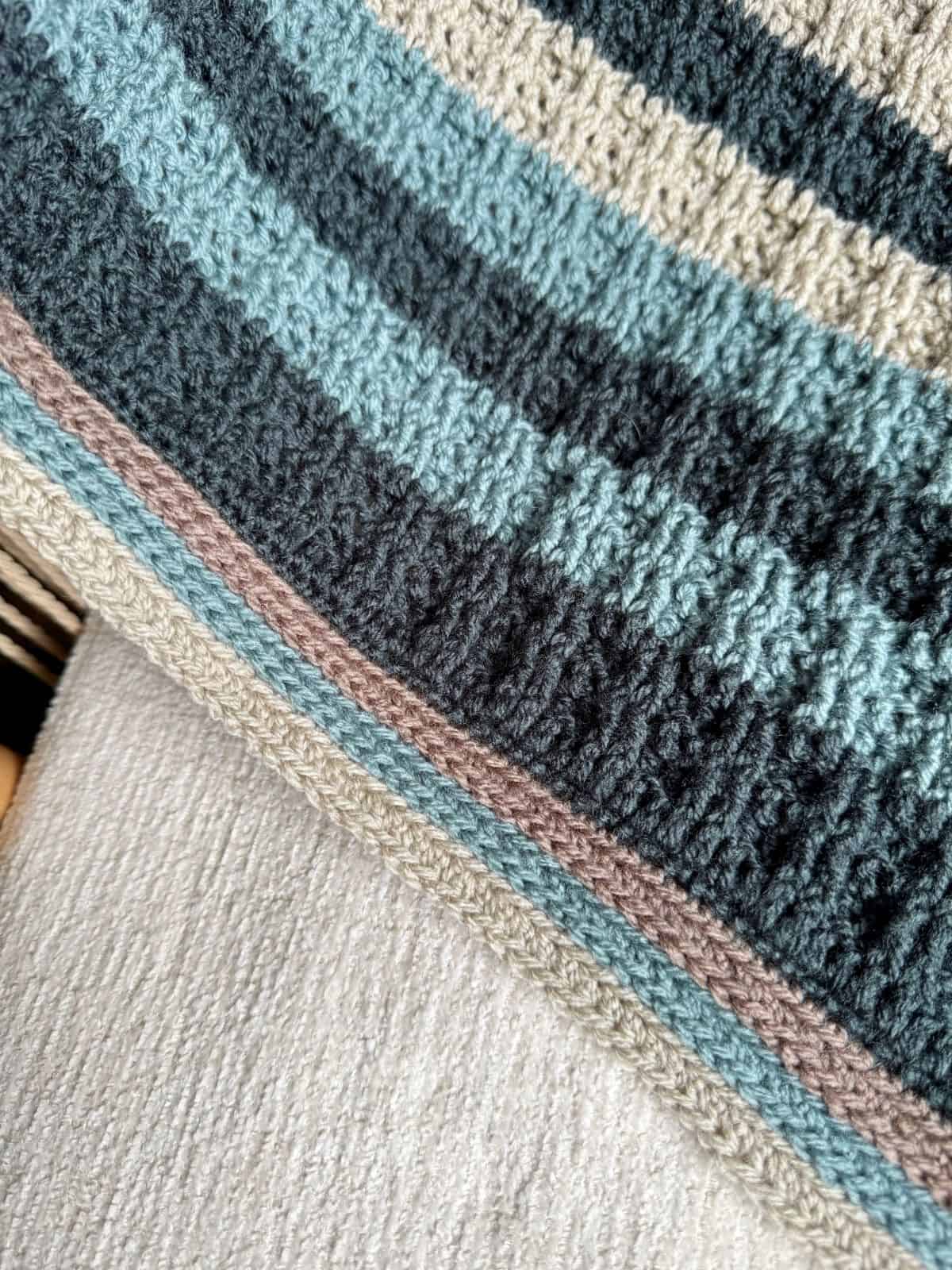 Close-up of a crochet blanket for boys featuring knitted horizontal stripes in shades of blue, gray, beige, and brown, draped over a textured light-colored surface.