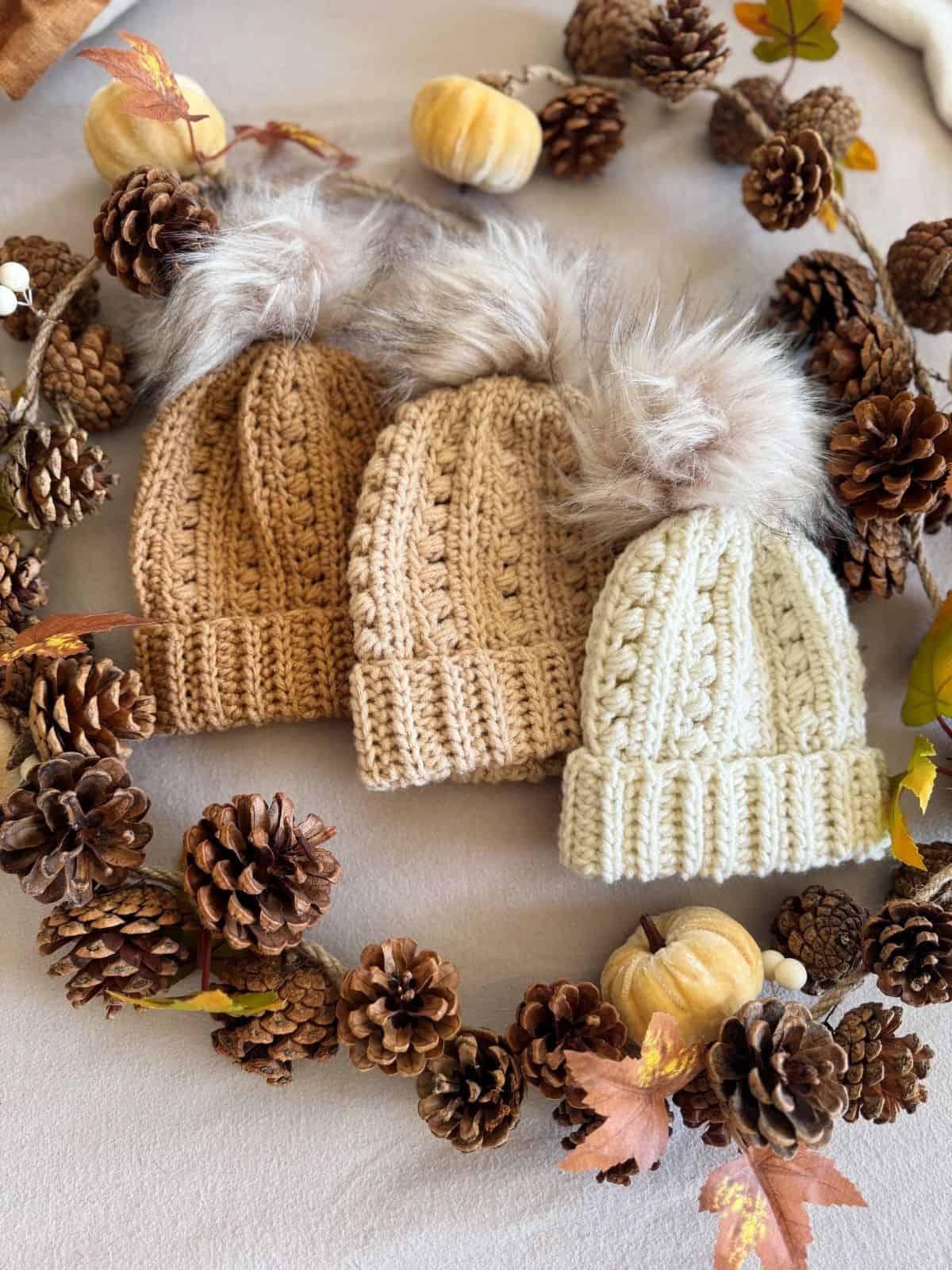Three crochet beanies with faux fur pom-poms, inspired by a puff stitch crochet hat pattern, are displayed on a table, surrounded by pinecones, autumn leaves, and small decorative pumpkins.