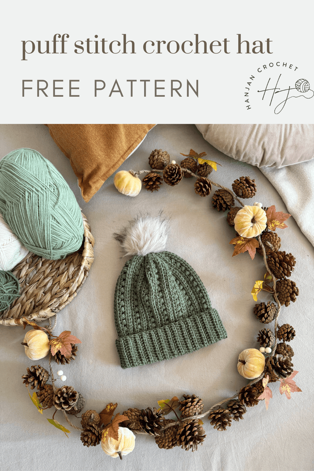 Flat lay of a green crocheted hat with a faux fur pom-pom, surrounded by yarn, pillows, and autumn decor, highlighting a cozy puff stitch crochet hat pattern free to try.