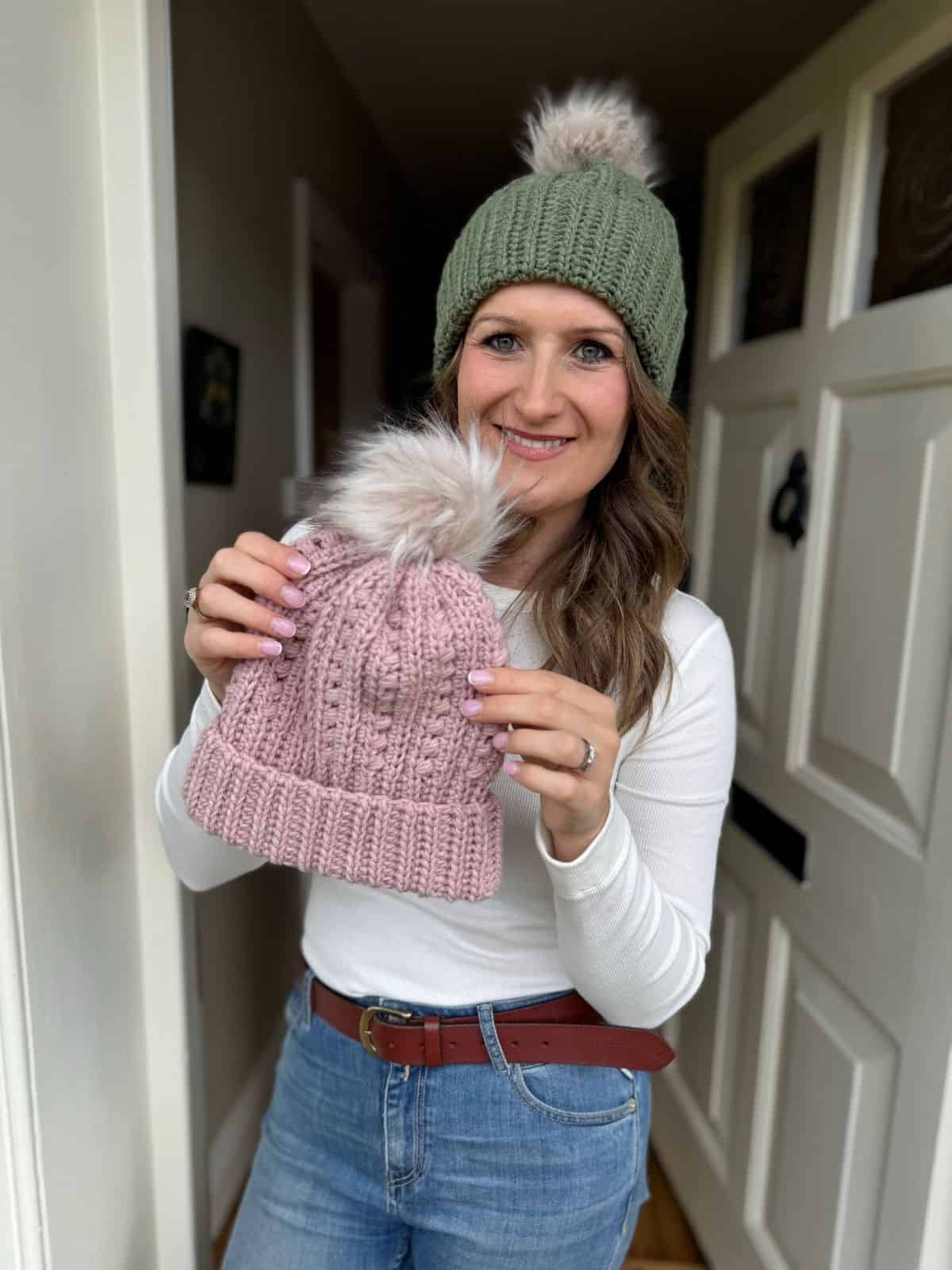 Woman in a white long-sleeve shirt and green knit hat stands indoors, holding up a pink puff stitch crochet hat with a fur pom-pom.