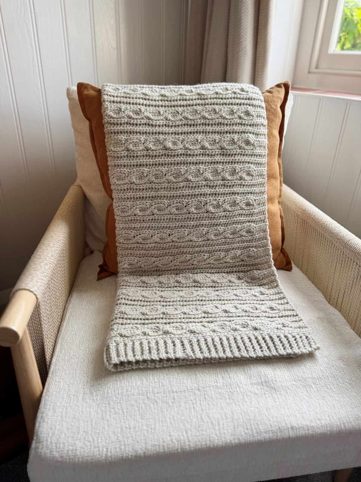 A textured, modern crochet blanket pattern in beige is draped over the back of a cushioned armchair, which sits beside a curtained window.