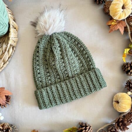A green puff stitch crochet hat pattern beanie with a white faux fur pom-pom on top, surrounded by pine cones, mini pumpkins, and autumn leaves.