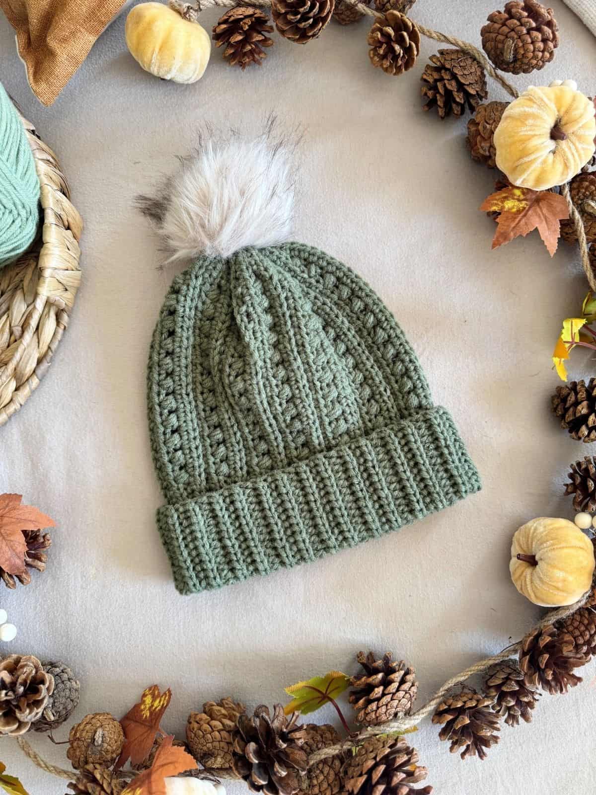 A green puff stitch crochet hat pattern beanie with a white faux fur pom-pom on top, surrounded by pine cones, mini pumpkins, and autumn leaves.