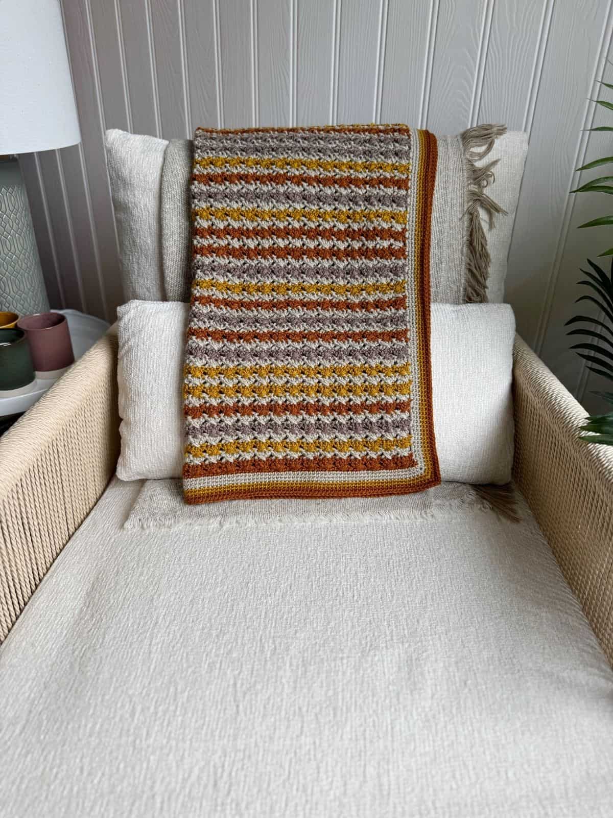 A crocheted, multicolored blanket with earth tones—crafted from a simple crochet blanket pattern—is draped over cream-colored cushions on a beige armchair beside a lamp and a plant.